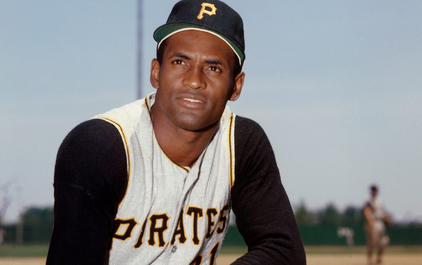 Roberto Clemente poses for a photo in his Pittsburgh Pirates uniform, outside on a baseball diamond.